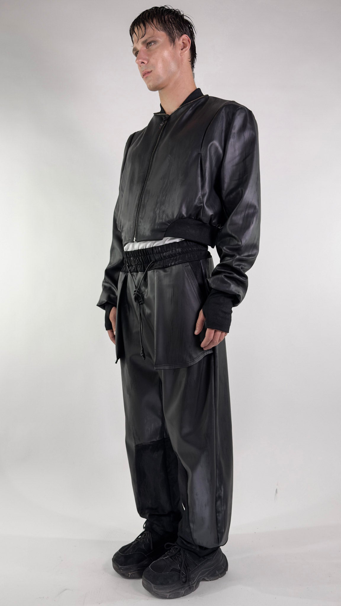 Pantalón Bomber de piel vegana negra con cintura elástica y cordón, junto a cazadora bomber corta; vista lateral